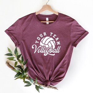 Puede incluir: Una camiseta color burdeos con un gr&aacute;fico de voleibol blanco y el texto "Your Team Volleyball" impreso en la parte delantera.