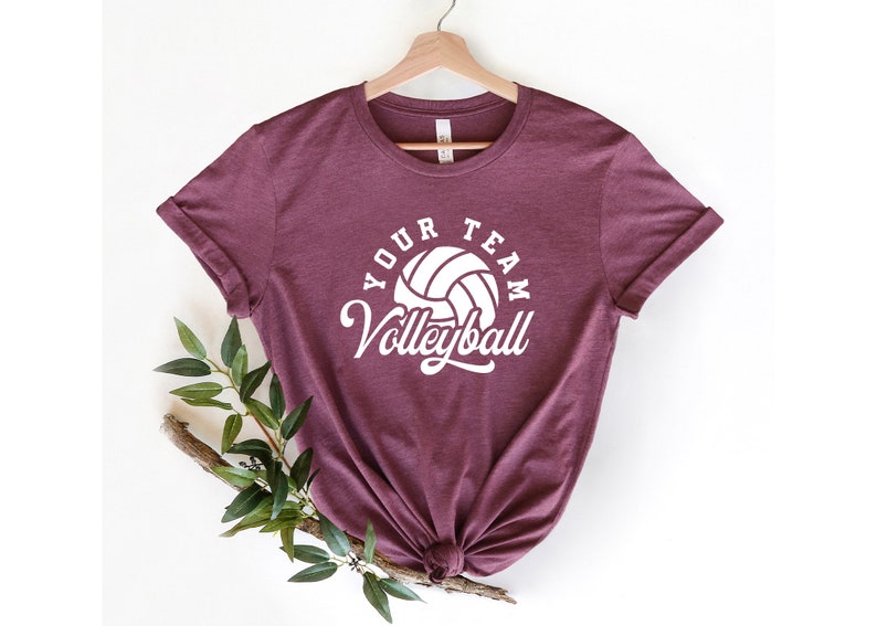 Puede incluir: Una camiseta color burdeos con un gr&aacute;fico de voleibol blanco y el texto "Your Team Volleyball" impreso en la parte delantera.