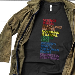May include: Black t-shirt with rainbow colored text that reads "Science is real, Black Lives Matter, No human is illegal, Love is love, Women's rights are human rights, Kindness is everything."