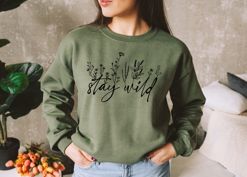 May include: A green sweatshirt with the text "stay wild" in black script with a line drawing of flowers and leaves.