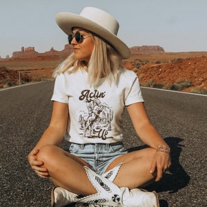 May include: A woman wearing a white cowboy hat, sunglasses, and a white t-shirt with a brown graphic that says "Actin' Up". She is sitting on a paved road with red rock formations in the background.
