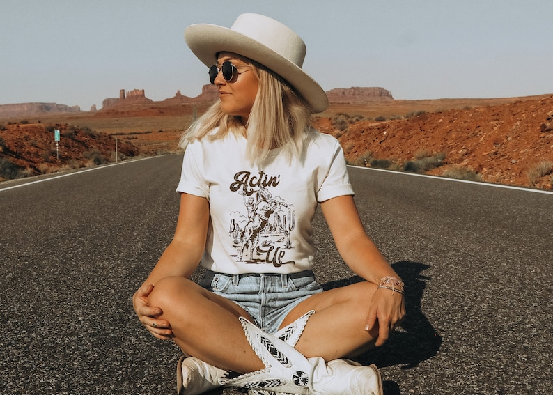 May include: A woman wearing a white cowboy hat, sunglasses, and a white t-shirt with a brown graphic that says "Actin' Up". She is sitting on a paved road with red rock formations in the background.
