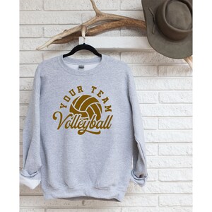 Puede incluir: Una sudadera gris con el texto "Your Team Volleyball" en letras marrones y un gr&aacute;fico de voleibol marr&oacute;n.