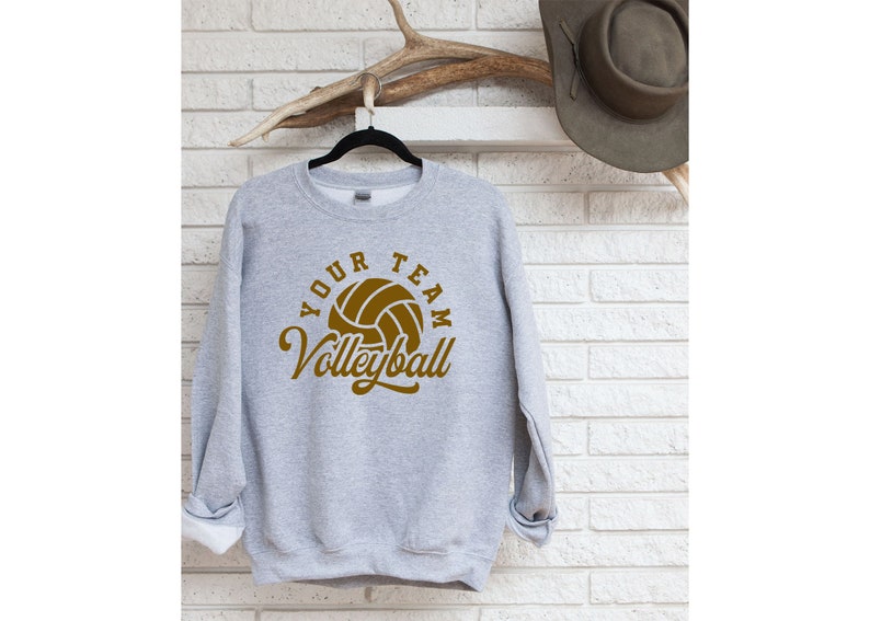 Puede incluir: Una sudadera gris con el texto "Your Team Volleyball" en letras marrones y un gr&aacute;fico de voleibol marr&oacute;n.