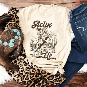 May include: A beige t-shirt featuring a brown graphic of a cowboy riding a horse in a desert landscape. The text "Actin' Up" is printed on the shirt.