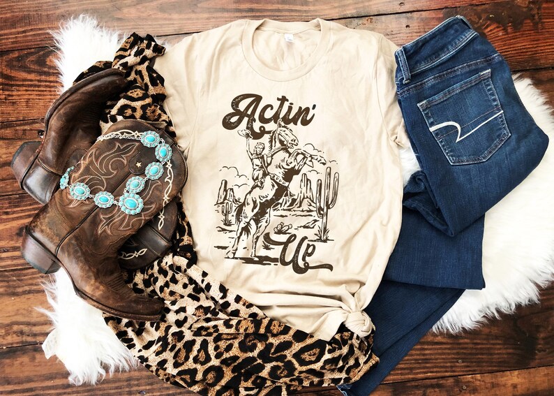 May include: A beige t-shirt featuring a brown graphic of a cowboy riding a horse in a desert landscape. The text "Actin' Up" is printed on the shirt.