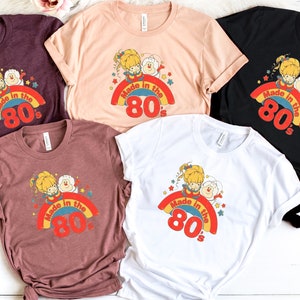 Puede incluir: Cinco camisetas de diferentes colores con un gr&aacute;fico de dibujos animados retro de dos figuras en un arco&iacute;ris con el texto "Made in the 80's".