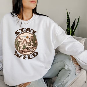May include: White sweatshirt with a brown graphic print of a desert landscape with cacti and the text "Stay Wild" in a circle.