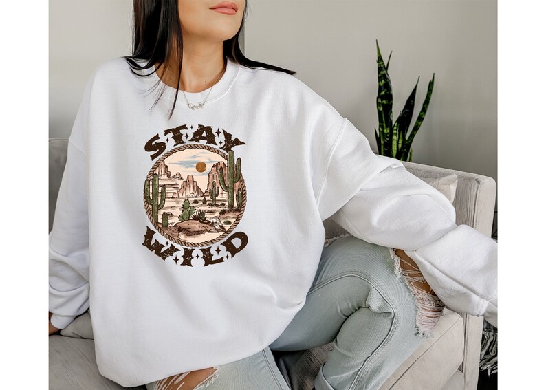 May include: White sweatshirt with a brown graphic print of a desert landscape with cacti and the text "Stay Wild" in a circle.