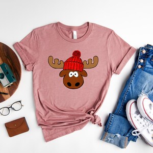May include: A mauve t-shirt with a cartoon moose wearing a red knit hat with a white pom-pom. The moose has brown antlers and a brown nose.