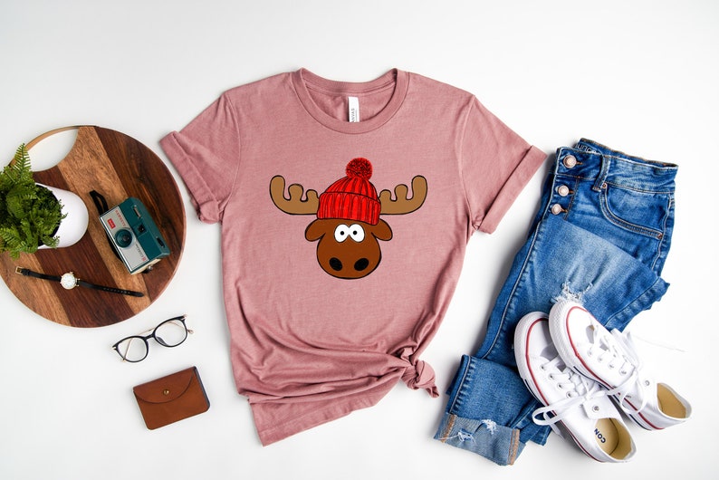 May include: A mauve t-shirt with a cartoon moose wearing a red knit hat with a white pom-pom. The moose has brown antlers and a brown nose.