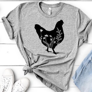 Pode incluir: Uma camiseta cinza com uma silhueta preta de uma galinha com flores dentro da silhueta. A camiseta est&aacute; colocada sobre uma superf&iacute;cie de madeira branca com um par de t&ecirc;nis brancos e um par de cal&ccedil;as jeans azuis.