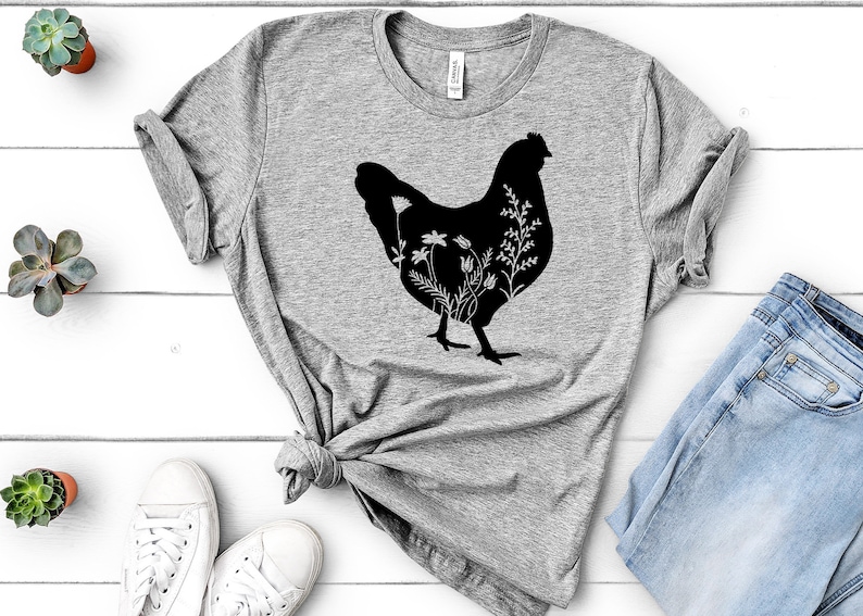 Pode incluir: Uma camiseta cinza com uma silhueta preta de uma galinha com flores dentro da silhueta. A camiseta est&aacute; colocada sobre uma superf&iacute;cie de madeira branca com um par de t&ecirc;nis brancos e um par de cal&ccedil;as jeans azuis.