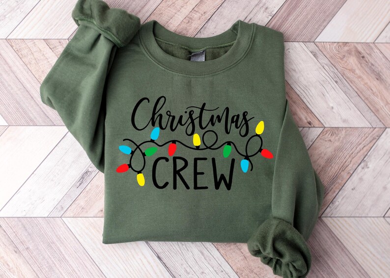 May include: A green sweatshirt with the text "Christmas Crew" in black lettering and a string of colorful Christmas lights.