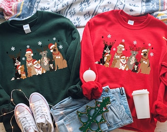 May include: Two Christmas sweaters with a green and red background featuring a group of dogs wearing Santa hats and reindeer antlers. The sweaters are laid out on a blue denim background with a white coffee mug and a Christmas tree ornament.