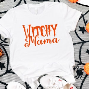 May include: White t-shirt with the orange text "Witchy Mama". The shirt is displayed on a black and white spiderweb-patterned surface, with small pumpkins and decorative spiders. A Halloween-themed fashion item.