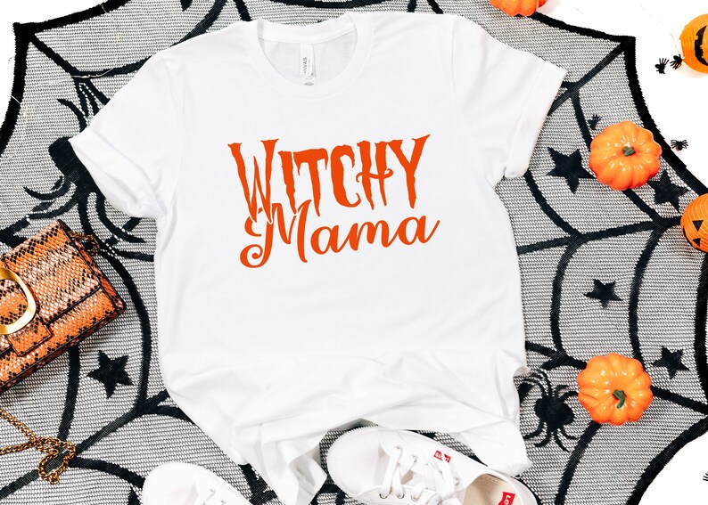 May include: White t-shirt with the orange text "Witchy Mama". The shirt is displayed on a black and white spiderweb-patterned surface, with small pumpkins and decorative spiders. A Halloween-themed fashion item.