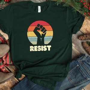 May include: A dark green t-shirt with a vintage-style graphic of a raised fist in front of a sunset. The text "RESIST" is printed below the graphic.