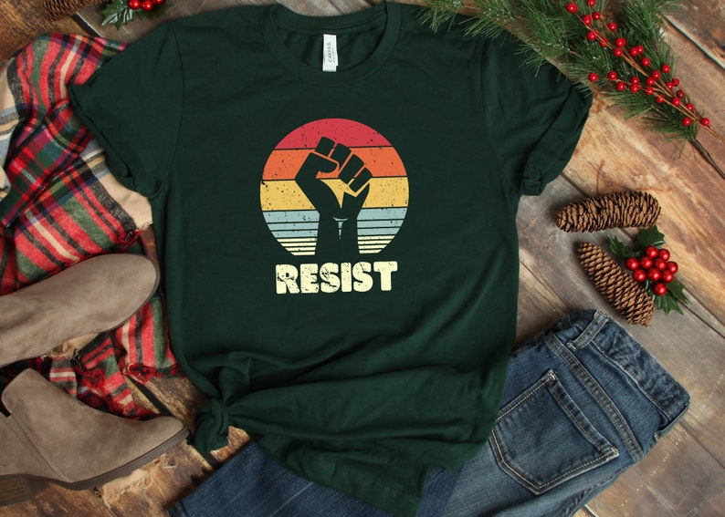 May include: A dark green t-shirt with a vintage-style graphic of a raised fist in front of a sunset. The text "RESIST" is printed below the graphic.