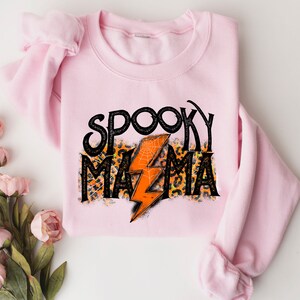 May include: A light pink sweatshirt with the words "Spooky Mama" in black, with an orange lightning bolt and leopard print accents. The sweatshirt is folded and displayed on a white surface, with floral decorations.