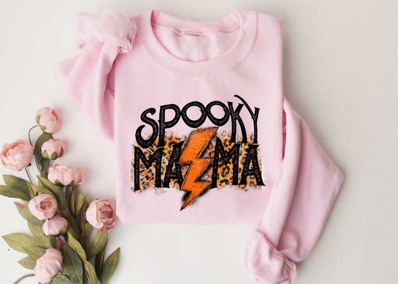 May include: A light pink sweatshirt with the words "Spooky Mama" in black, with an orange lightning bolt and leopard print accents. The sweatshirt is folded and displayed on a white surface, with floral decorations.