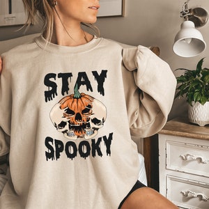 以下が含まれることがあります： ハロウィーンをテーマにしたベージュのスウェットシャツ。デザインは、スカルと黒い文字「STAY SPOOKY」が付いたジャック・オー・ランタンが特徴です。スウェットシャツは長袖で、ゆったりとしたフィット感です。