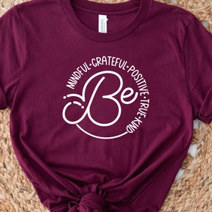 May include: A maroon t-shirt with a white circle design that says "Be" with the words "Mindful, Grateful, Positive, True, Kind" surrounding the word "Be".