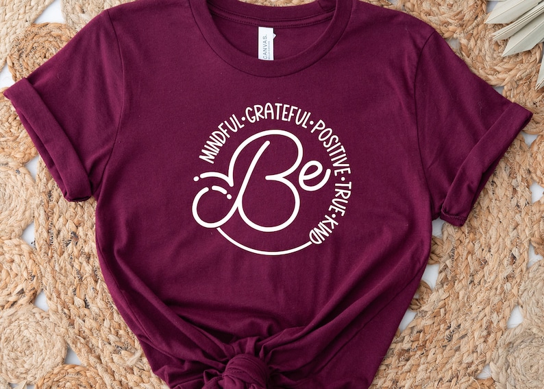 May include: A maroon t-shirt with a white circle design that says "Be" with the words "Mindful, Grateful, Positive, True, Kind" surrounding the word "Be".