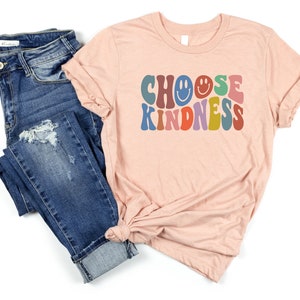 May include: A peach-colored t-shirt with the text "CHOOSE KINDNESS" in colorful retro font. The text includes a smiley face. The shirt is paired with blue jeans.