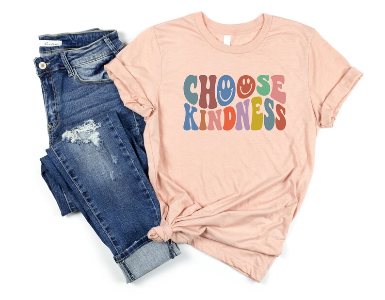 May include: A peach-colored t-shirt with the text "CHOOSE KINDNESS" in colorful retro font. The text includes a smiley face. The shirt is paired with blue jeans.