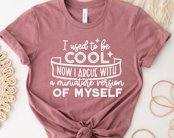 May include: A mauve t-shirt with white text that reads "I used to be COOL NOW I ARGUE WITH a miniature version OF MYSELF".