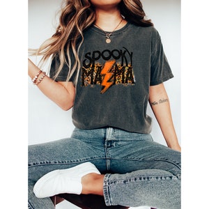 May include: Dark gray t-shirt with the text "SPOOKY MAMA" in a black, leopard print, and orange lightning bolt design. The shirt is a casual, comfortable style, perfect for Halloween or spooky season.