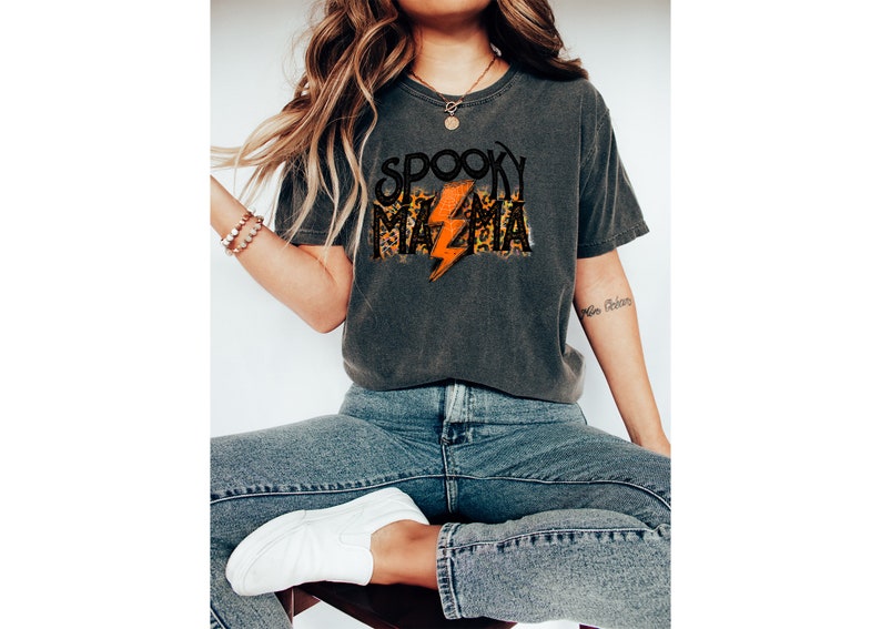 May include: Dark gray t-shirt with the text "SPOOKY MAMA" in a black, leopard print, and orange lightning bolt design. The shirt is a casual, comfortable style, perfect for Halloween or spooky season.