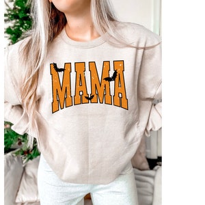 May include: Beige sweatshirt with the word "MAMA" in orange block letters with a black outline. Three black bats are above and between the letters. The sweatshirt is a Halloween-themed design.