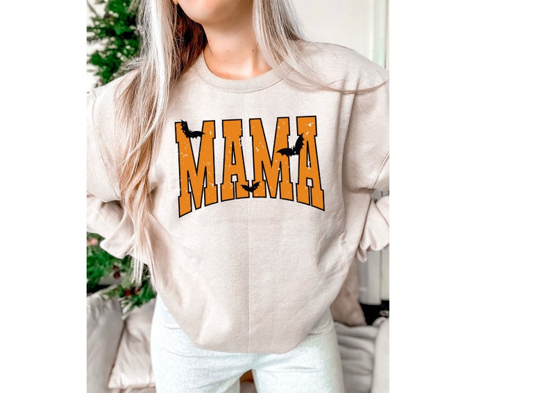 May include: Beige sweatshirt with the word "MAMA" in orange block letters with a black outline. Three black bats are above and between the letters. The sweatshirt is a Halloween-themed design.