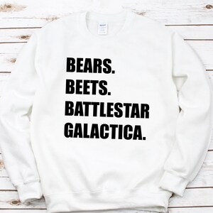 May include: White sweatshirt with the text "BEARS. BEETS. BATTLESTAR GALACTICA." in bold black letters. The sweatshirt is a crew neck style and appears to be made of a soft material. The background is a light wood grain.