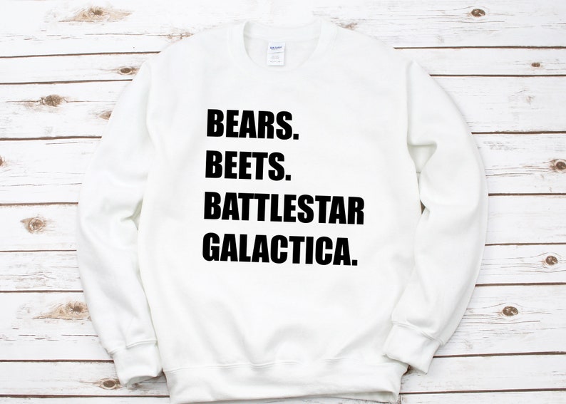 May include: White sweatshirt with the text "BEARS. BEETS. BATTLESTAR GALACTICA." in bold black letters. The sweatshirt is a crew neck style and appears to be made of a soft material. The background is a light wood grain.