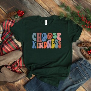 May include: A dark green t-shirt with the text "Choose Kindness" in rainbow colors with smiley faces. The text is in a retro font style.
