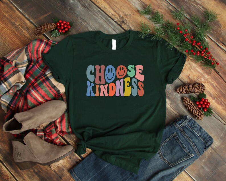 May include: A dark green t-shirt with the text "Choose Kindness" in rainbow colors with smiley faces. The text is in a retro font style.