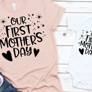 May include: Two shirts with the text "Our First Mother's Day" in black lettering with stars and hearts. The larger shirt is pink and the smaller shirt is white.