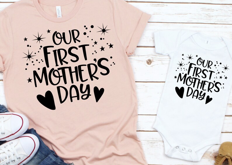 May include: Two shirts with the text "Our First Mother's Day" in black lettering with stars and hearts. The larger shirt is pink and the smaller shirt is white.