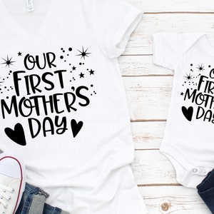 May include: Two white t-shirts with black text and hearts. The larger shirt says "Our First Mother's Day" with stars and hearts. The smaller shirt says "Our First Mother's Day" with stars and hearts.