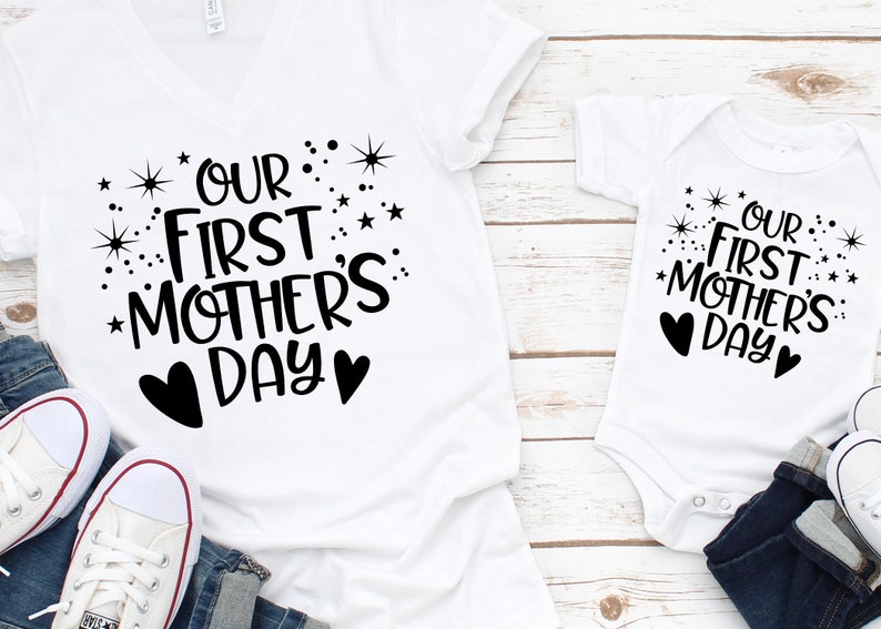 May include: Two white t-shirts with black text and hearts. The larger shirt says "Our First Mother's Day" with stars and hearts. The smaller shirt says "Our First Mother's Day" with stars and hearts.
