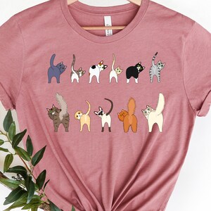 May include: A pink t-shirt with a graphic of multiple cats with their tails up in the air. The cats are all different colors and breeds.
