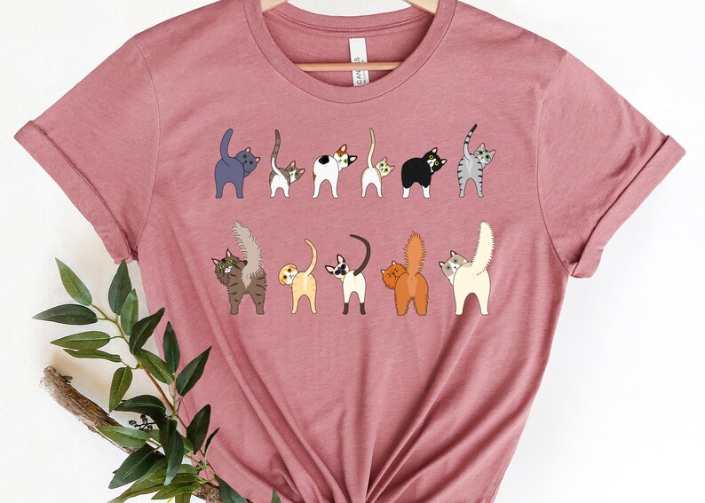 May include: A pink t-shirt with a graphic of multiple cats with their tails up in the air. The cats are all different colors and breeds.