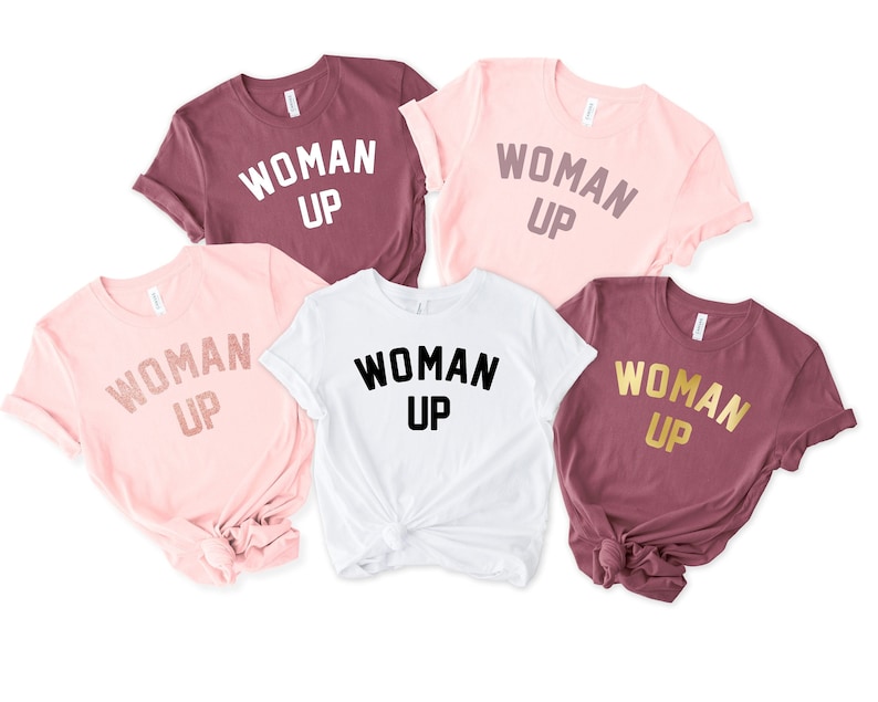 May include: Five t-shirts with the text "WOMAN UP" printed on them. The shirts are in different colors: maroon, pink, white, and two shades of pink. The text is in different colors: white, gold, and black.