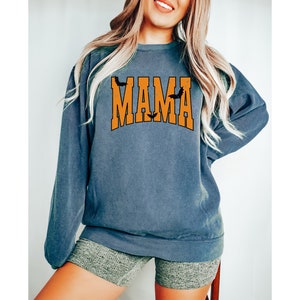 May include: A blue-grey sweatshirt with the word "MAMA" in orange block letters. The word "MAMA" has a bat on top of each letter "A". The sweatshirt is a crew neck style.