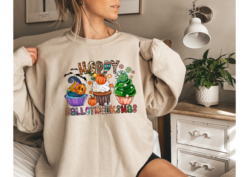 K&ouml;nnte beinhalten: Beiges Sweatshirt mit einem farbenfrohen Aufdruck mit der Aufschrift "HAPPY HALLOTHANKSMAS". Das Design zeigt Cupcakes, die mit Halloween- und Thanksgiving-Themen dekoriert sind, darunter K&uuml;rbisse, Flederm&auml;use und Schneeflocken. Ein gem&uuml;tliches, festliches Oberteil.