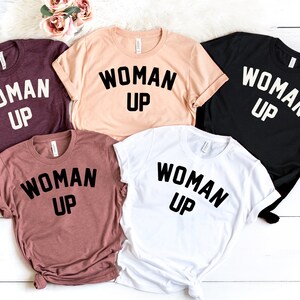 May include: Five t-shirts in different colors with the text "WOMAN UP" printed in black on each shirt.