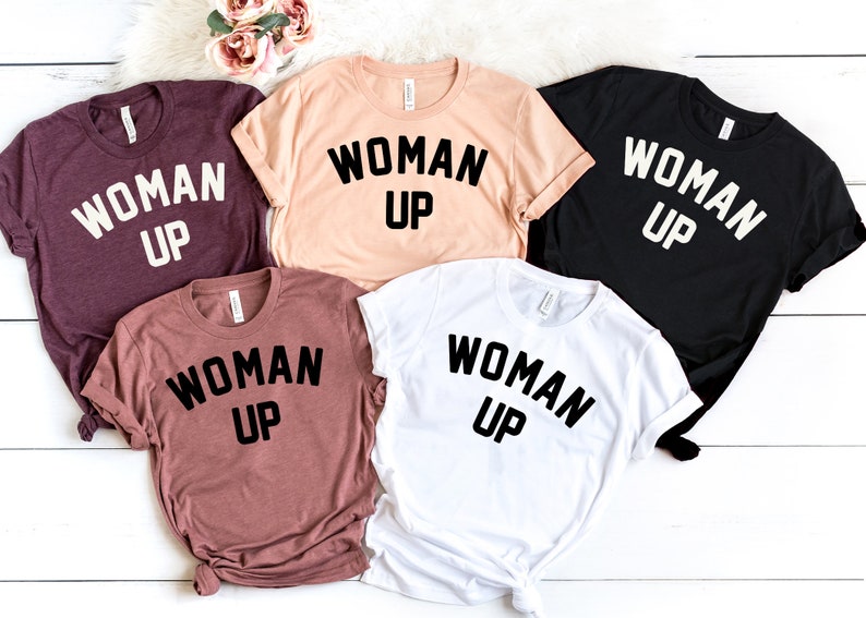 May include: Five t-shirts in different colors with the text "WOMAN UP" printed in black on each shirt.